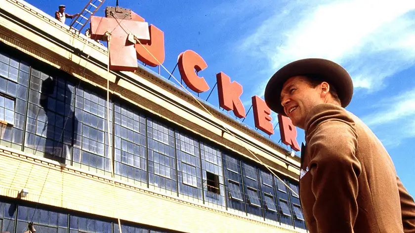 A man stands smiling wearing a hat and a brown suit in front of the TUCKER department store as someone is putting up the letter T of the logo on the roof