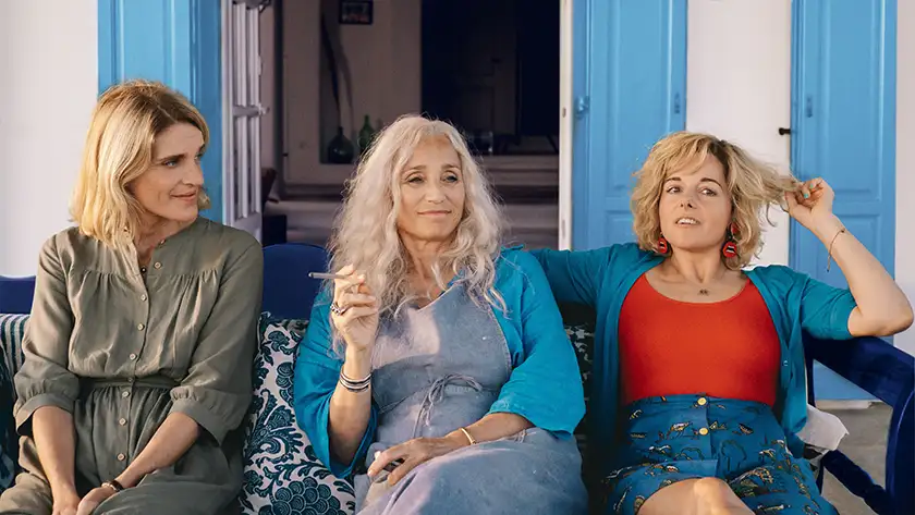 Three middle aged women sit on a blue outdoors sofa, the one in the middle smoking a cigarette, in front of a white wall with light blue windows in the film Two Tickets to Greece