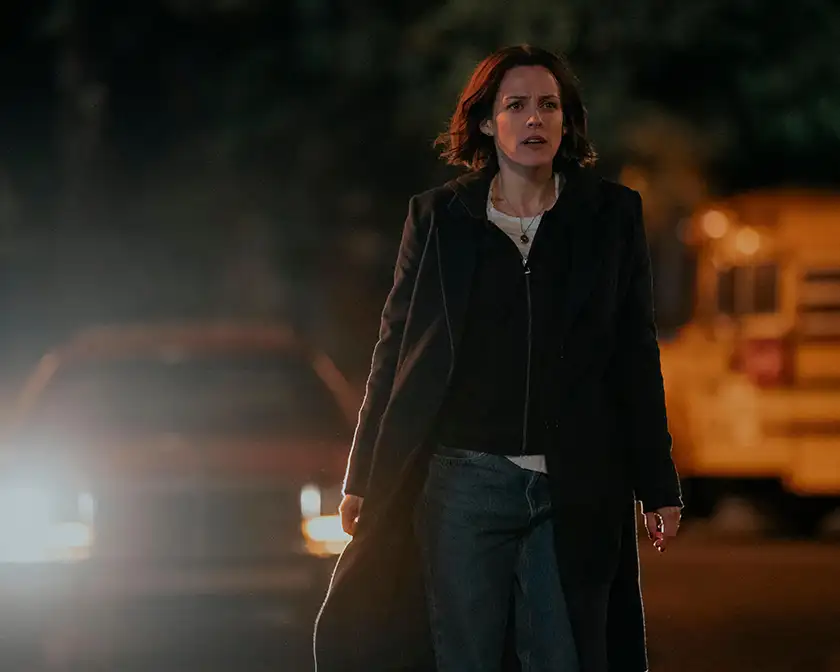 A woman stands in the middle of the street at night with a car behind her, looking worried, in Episode 6 of Under the Bridge