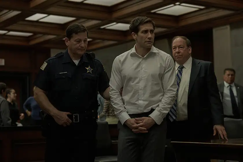 A man with a white shirt is being taken to his seat by a police officer in a courtroom in the Apple TV+ series Presumed Innocent
