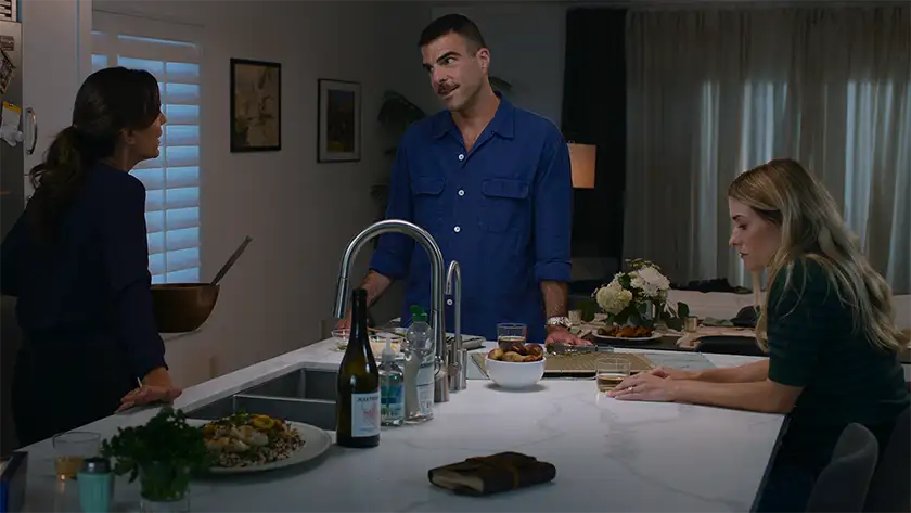 Three characters are around a dinner table, two standing and one sitting, in the film Adult Best Friend, in a photo featured on the Loud and Clear interview with Delaney Buffett and Katie Corwin