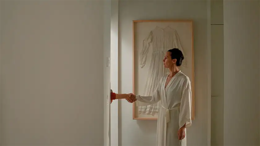 A woman in a white bathrobe shakes someone's hand, but the hand's owner is hidden behind a white wall, in the film Family Therapy
