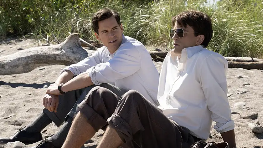 Two men wearing white shirts sit on the beach in Fellow Travelers, in a photo featured in an article about why Tim's conflict between faith and sexuality matters in the series