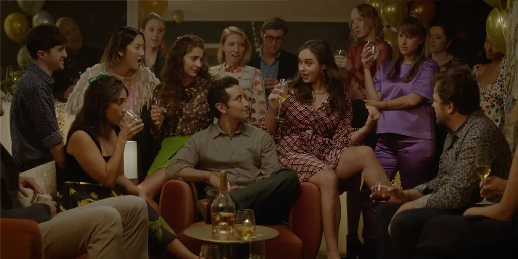 A man sits by a table surrounded by happy people in a still from the 2024 film The French Italian
