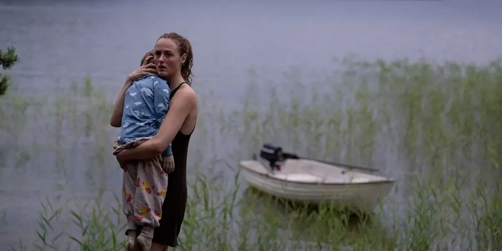 A woman stands holding a child in her hands with a boat behind her in shock in a still from the film Handling the Undead