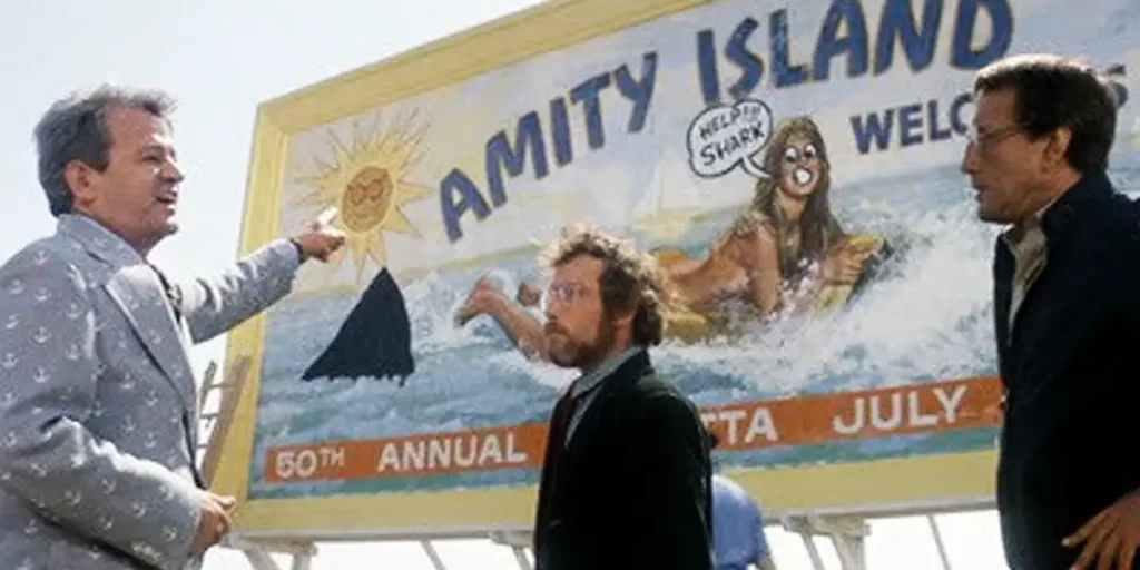 A man points at a sign that reads Amity Island with two other men looking at him in a still from Jaws, the perfect fourth of july movie