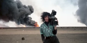 War photographer Margaret Moth holds a videocamera with an explosion behind her in a still from the documentary film Never Look Away