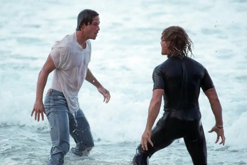 Keanu Reeves and Patrick Swayze are in the water, standing opposite each other with wet clothes, in the film Point Break