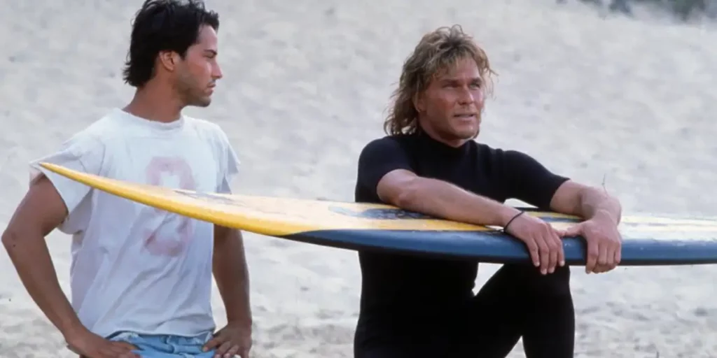 Keanu Reeves and Patrick Swayze sit on the beach, the latter holding a surf board, in the film Point Break