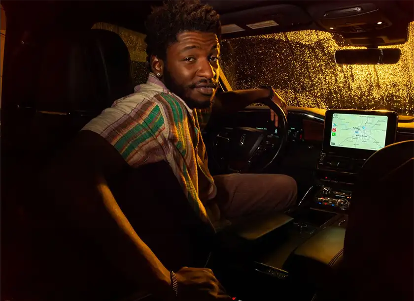 A Black man sits in a car with a navigating system turned on, looking back at the camera and smiling, in the film Rob Peace