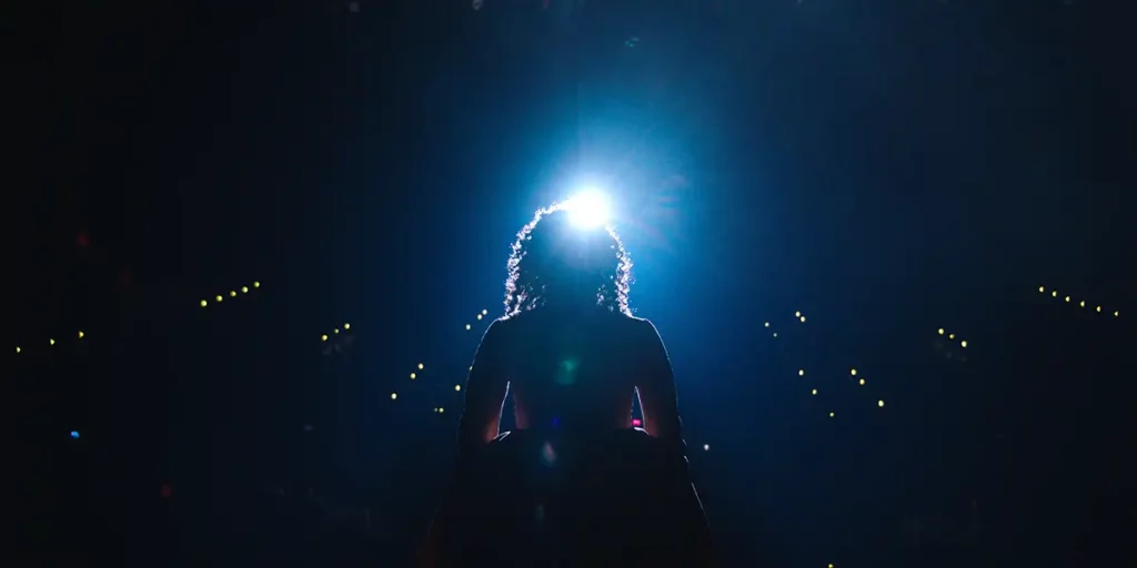 An outlet of someone on the stage faces the audience with a light in front of them in the dark in a still from the 2024 film Satisfied
