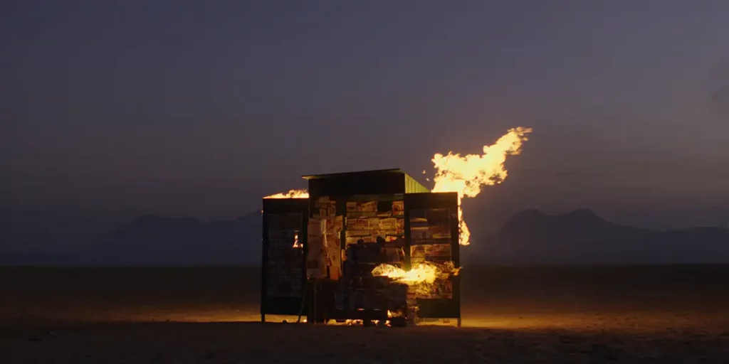 A wooden structure with magazines on them is on fire at night in a still from the film State of Silence