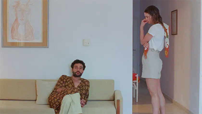 A woman stands, looking at a man sitting on the sofa, in a still from the film Swimming Home
