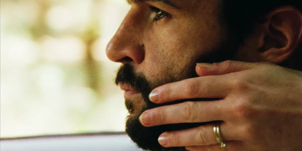 A man's face with hands touching his beard in a still from the film Swimming Home