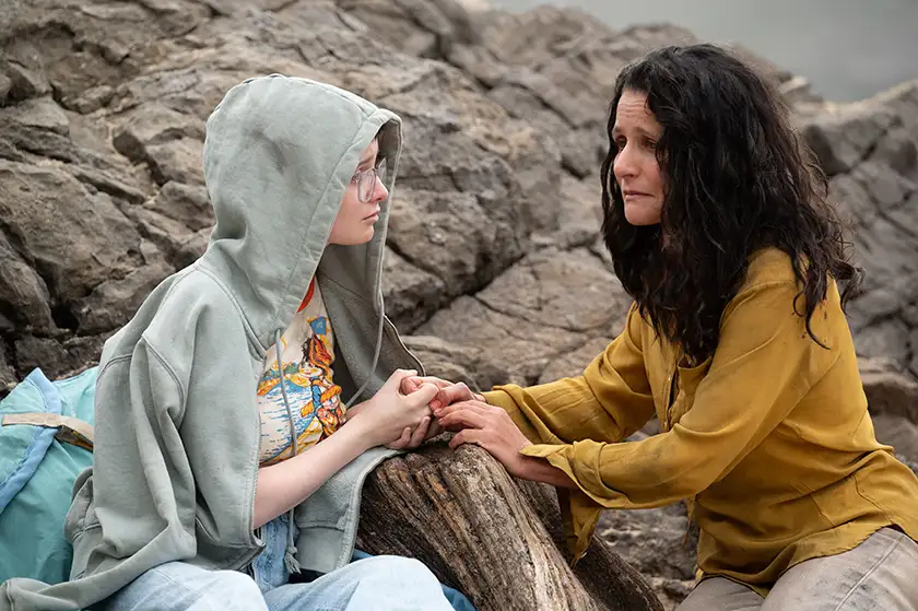 A mother holds a daughter's hands and they are both crying while leaning on some rocks in a still from the film Tuesday