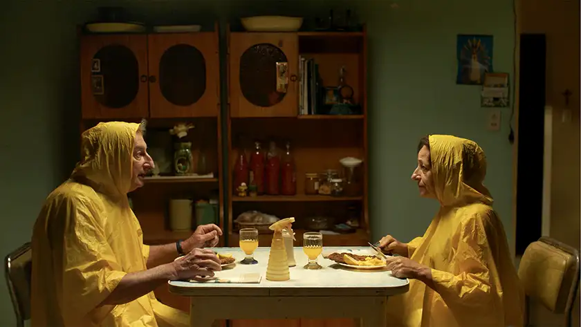 Husband and wide sit at the dinner table wearing bright yellow raincoats and drinking yellow water in the film Chronicles of a Wandering Saint