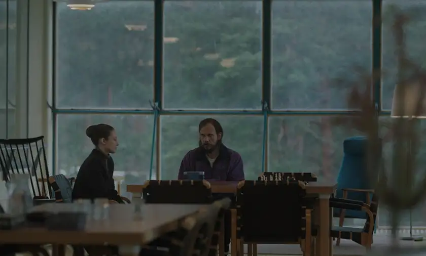 A man with a purple shirt, long hair and a beard and a woman with brown hair sit at a table looking sad in the film What Remains