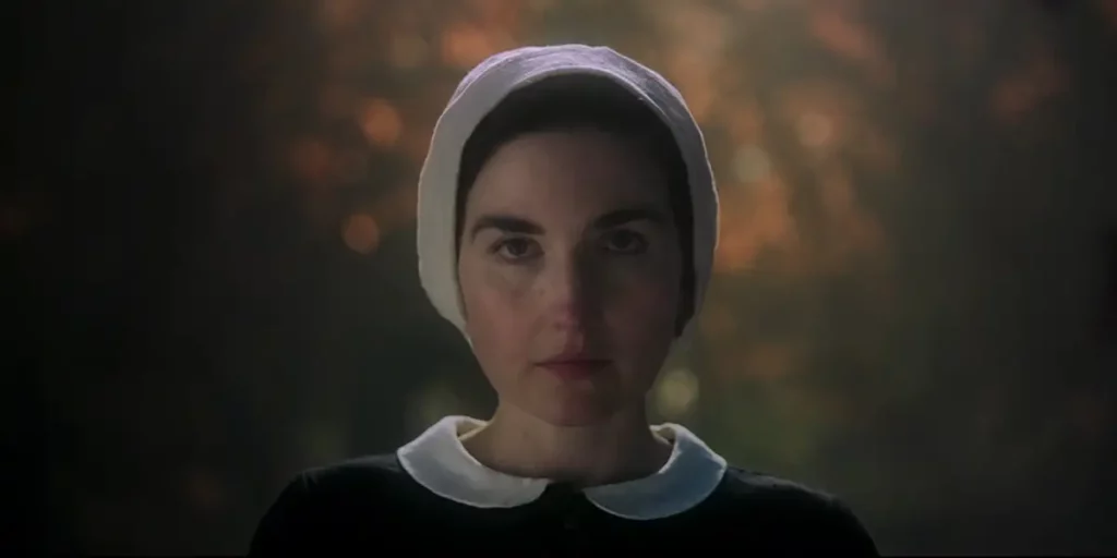 A young woman with a white cloth on her head and a black dress stands looking at the camera with fire behind her in a still from the 2024 film Witches
