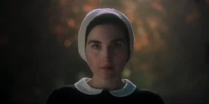 A young woman with a white cloth on her head and a black dress stands looking at the camera with fire behind her in a still from the 2024 film Witches