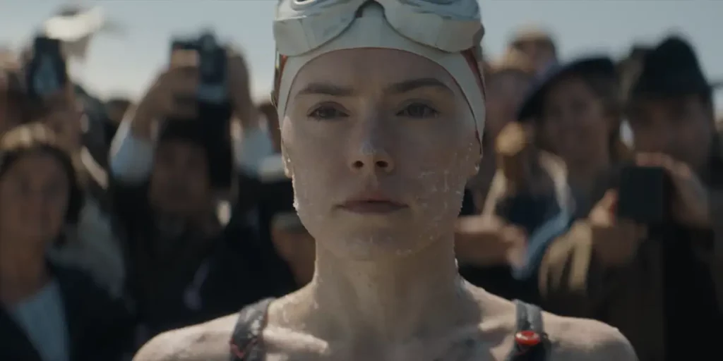 A woman wears a swimming cap and looks ahead surrounded by a cheering crowd in the film Young Woman and the Sea