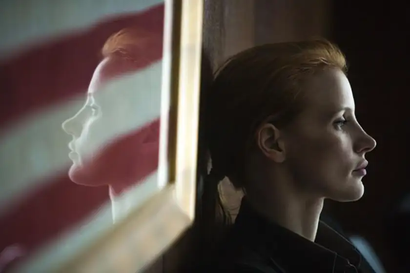 A red haired woman with a ponytail leans against a wall next to a framed US flag in the film Zero Dark Thirty