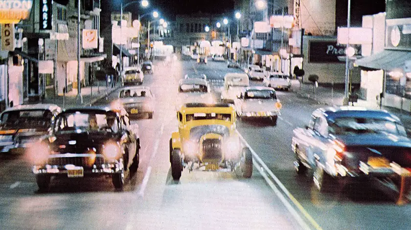 A yellow car is going against the flow on a road at night in the film American Graffiti