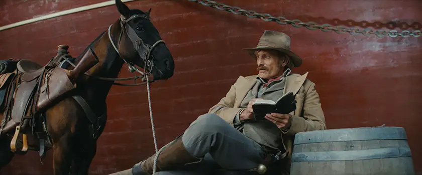 Viggo Mortensen leans on a wall reading a book with a saddled horse next to him in The Dead Don't Hurt