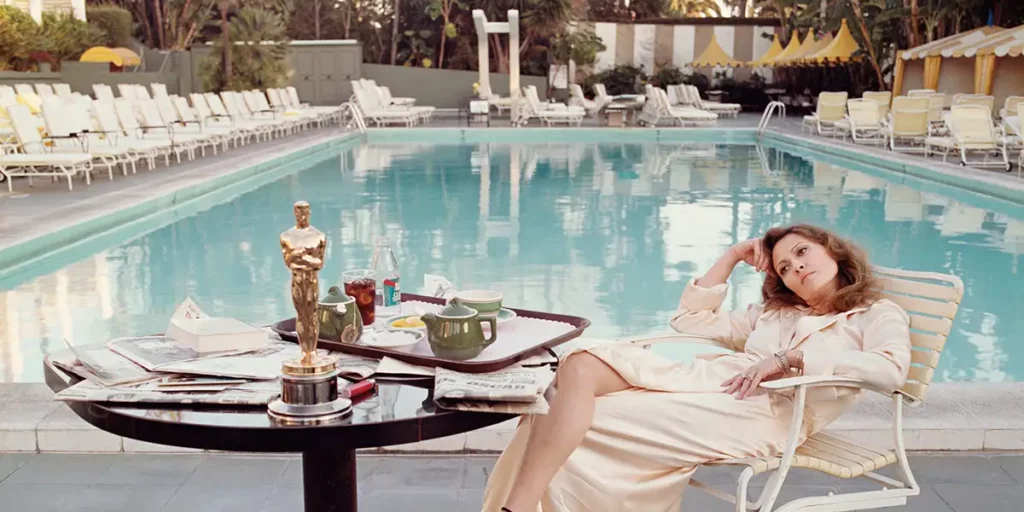 Faye Dunaway lies on a white chair by. the swimming pool with her Oscar on a table in front of her next to a tray with her breakfast, and newspapers underneath both, in the main still for the 2024 HBO documentary "Faye"