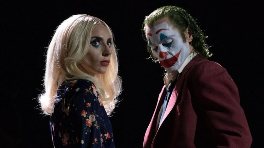 Lady Gaga and Joaquin Phoenix stand close to one another dressed as Harley Quinn and Joker against a black background in Folie à Deux