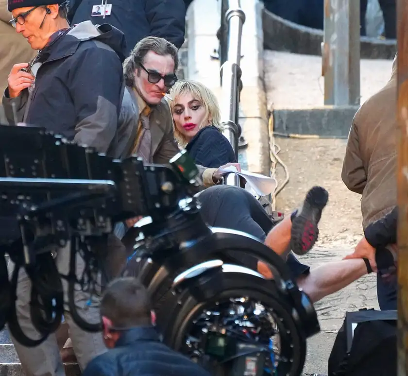 Lady Gaga and Joaquin Phoenix sit with smudged makeup in a set photo of Joker 2: Folie à Deux, featured in a Loud and Clear article with everything we know about the movie