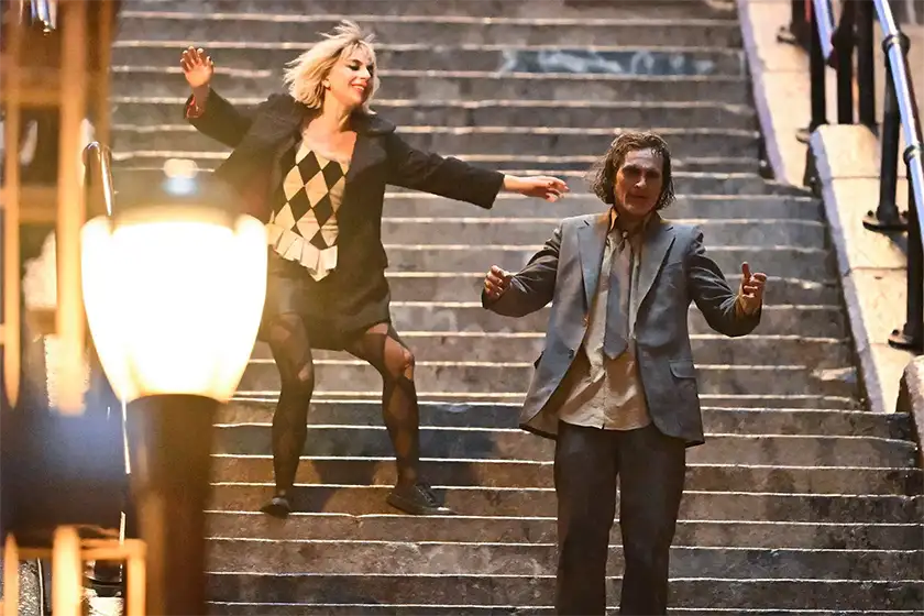 Lady Gaga and Joaquin Phoenix dance on the stairs in a set photo of Joker 2: Folie à Deux, featured in a Loud and Clear article with everything we know about the movie