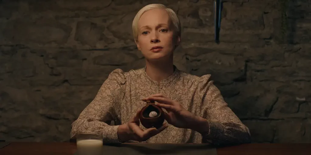 A blonde woman holds a compass in her hands while sitting in a still from the supernatural whodunit horror film ODDITY