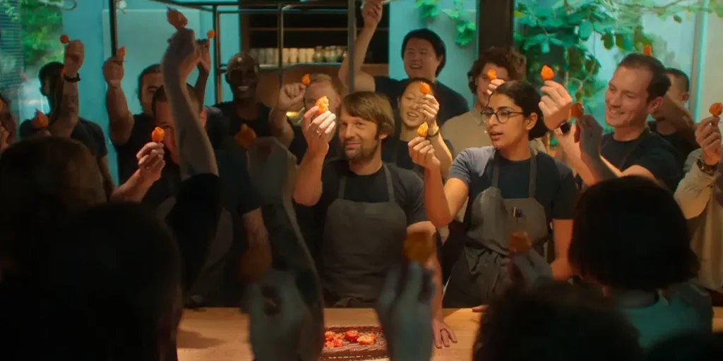 A chef holds an ingredient surrounded by many people in a still from the Apple TV+. series Omnivore