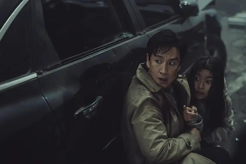 Two scared people lean against a car looking to their right in the film Project Silence