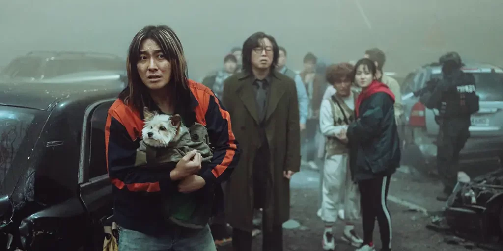 A woman with a red and black jacket holds a small white dog and looks ahead, with people and wreckage behind her, in the film Project Silence