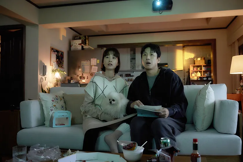 An Asian couple sits on a sofa looking at something in fear, holding a small dog and some papers, in Jason Yu's film Sleep