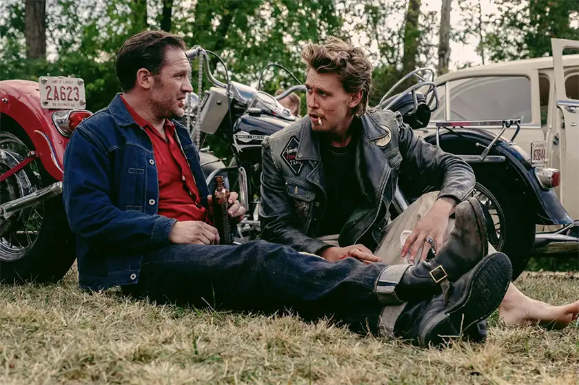 Two bikers sit on the grass talking and looking at each other in the film The Bikeriders