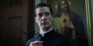 A priest stands, looking down, with a religious painting behind him in the 2024 film Ababouiné