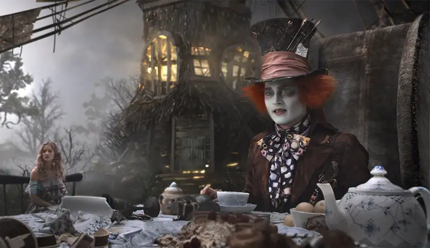 The made hatter sits at a table with a cup in front of him and Alice sitting next to him, much smaller than normal, in the film Alice in Wonderland (2010)