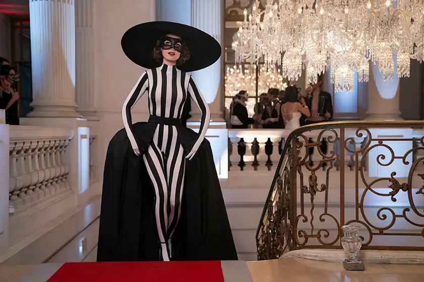 Lily Collins walks up the stairs with a big black hat, a black mask and a striped dress at a masquerade ball as Emily in Season 4 Part 1 (episode 4) of Emily in Paris