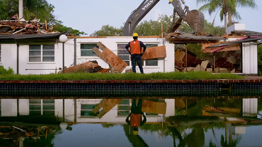 A construction worker stands looking at a house being destructed, with his outline reflected on water, in the film Mountains