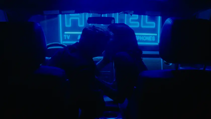 Two people kiss in a car with a neon blue sign in front of them, in a shot seen from the back of the car, in the film Strange Darling