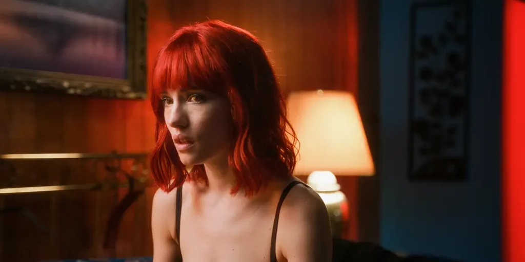 A red haired woman wears a black tanktop and sits with a lamp behind her in the film Strange Darling