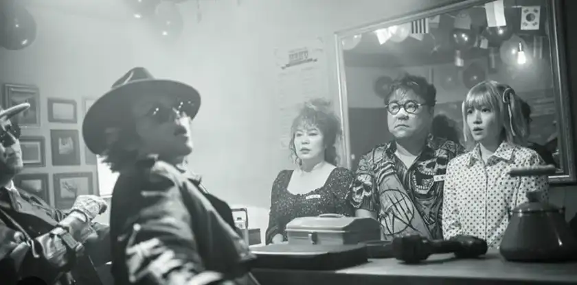 Three people look ahead when another person, sitting opposite them at a table, looks at the camera in a black and white still from Silent CInema, one of the shorts from the 2024 crime anthology The Killers