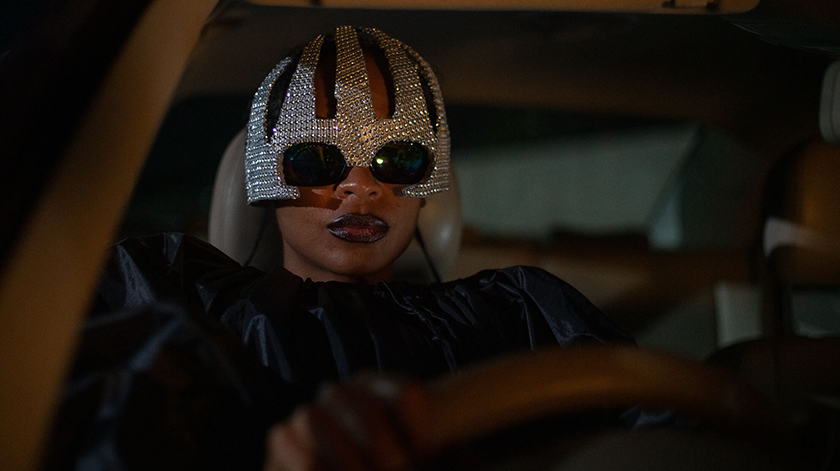 Susan Chardy, as Schula, drives a car at night, wearing sunglasses and a sparkly hat but with a stern expression, in On Becoming a Guinea Fowl