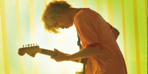 A Japanese boy plays a guitar standing by a yellow curtain and wearing an orange t-shirt in the film Plastic