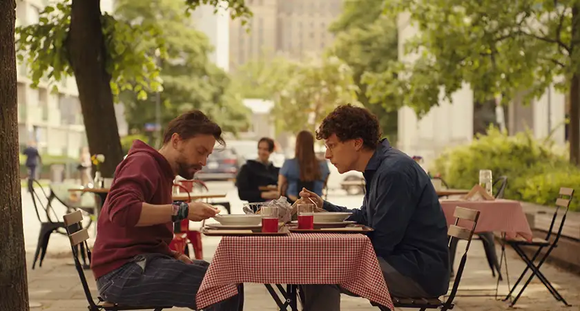 Kieran Culkin and Jesse Eisenberg sit across each other at a table while having lunch outdoors in the film A Real Pain, which cements Jesse Eisenberg’s creative focus according to Loud and Clear Reviews