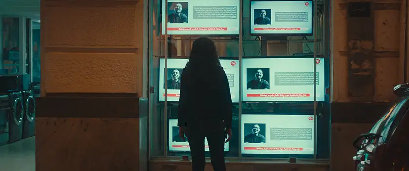 A woman looks at TV screens in a shop window at night in a still from the movie Aïcha, whose director, Mehdi Barsaoui, we interview