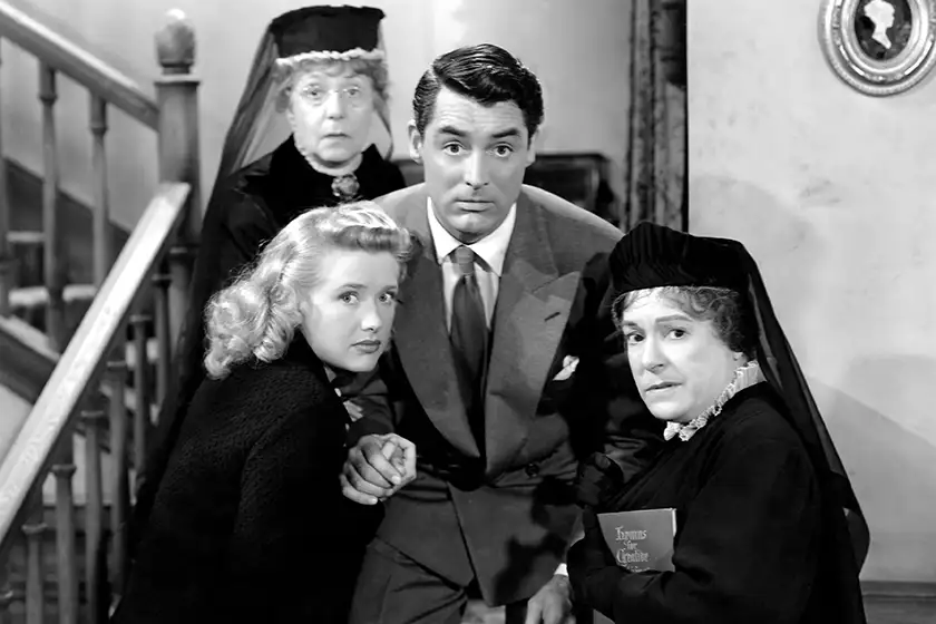 Cary Grant and three women are on a staircase in a black and white still from Arsenic and Old Lace, one of the 10+ movies to watch at Halloween 2024 according to Loud and Clear Reviews