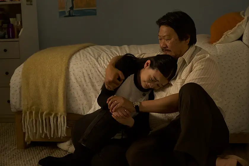 Two characters hug each other sitting by a bed in a still from the film Bad Genius